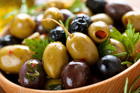 Bowl of assorted gourmet Mediterranean olives including green Castelvetrano, black Kalamata, and herb-marinated varieties on rustic wood table