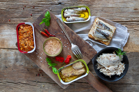 Assorted gourmet anchovy tins and jars from Italy and Spain on a rustic wooden table, ideal for pasta, pizza, and antipasti dishes